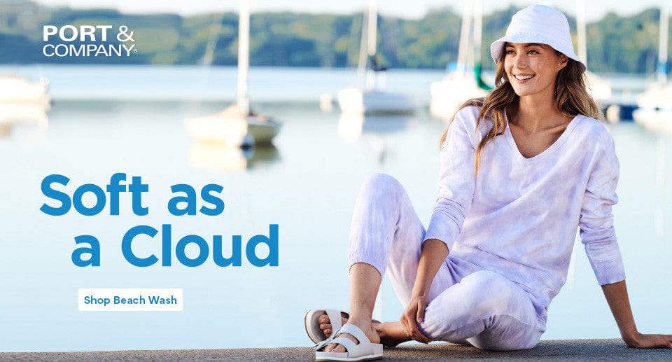 Woman in white outfit sitting by a waterfront with 'Port & Company' branding and text 'Soft as a Cloud'.