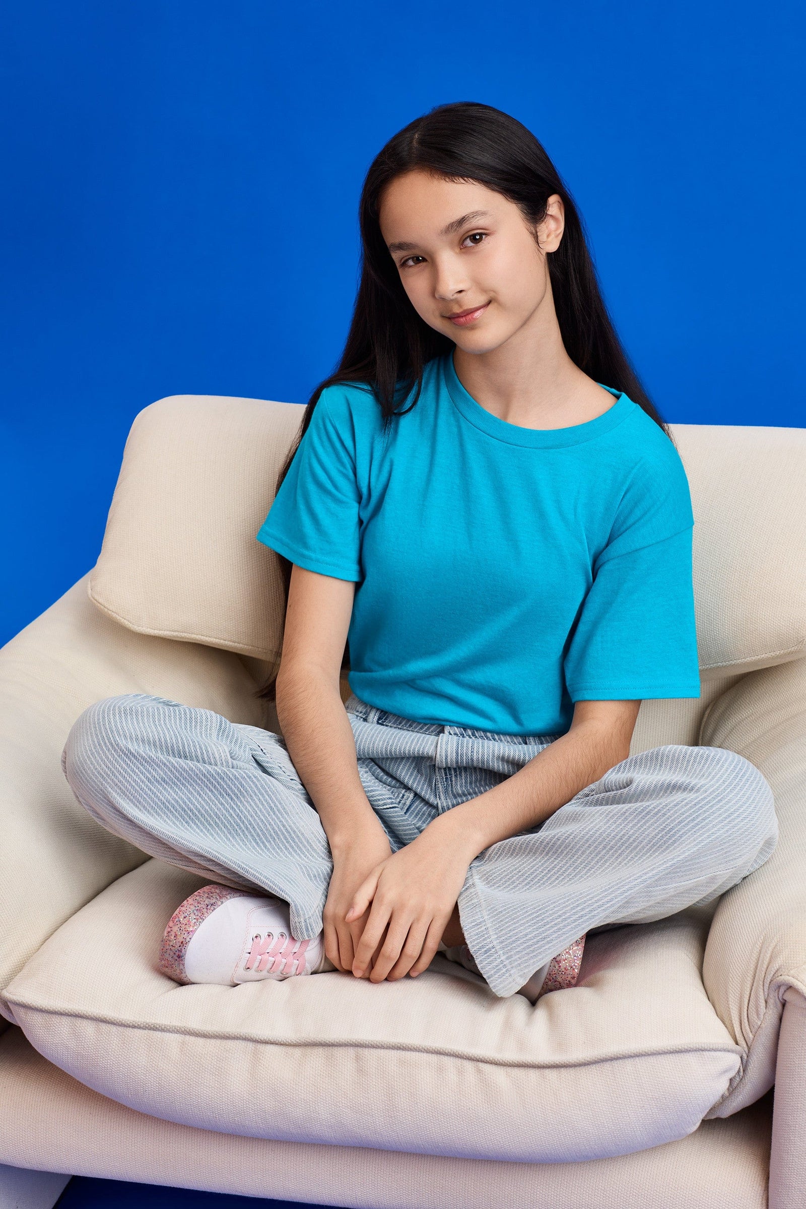 Young girl sitting on a beige couch wearing a blue shirt and light gray pants against a blue background