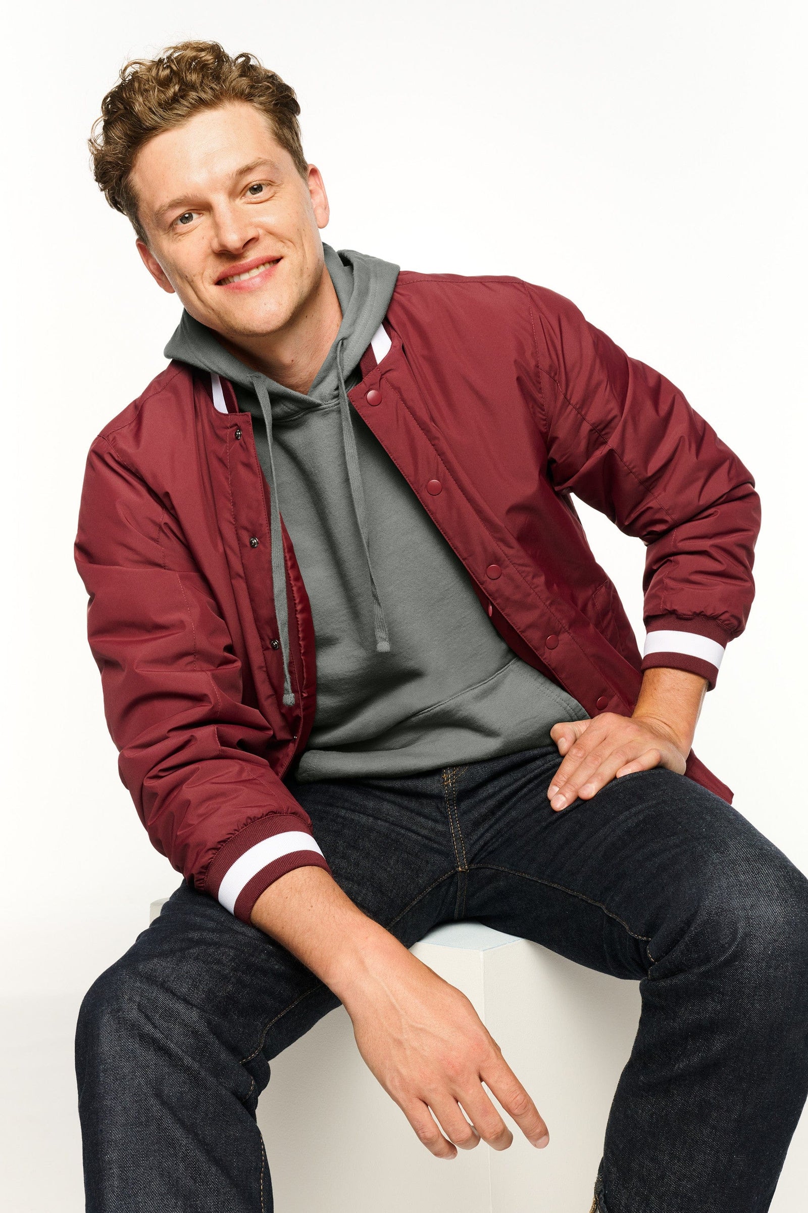 Man wearing a maroon varsity jacket over a gray hoodie, sitting on a white chair.